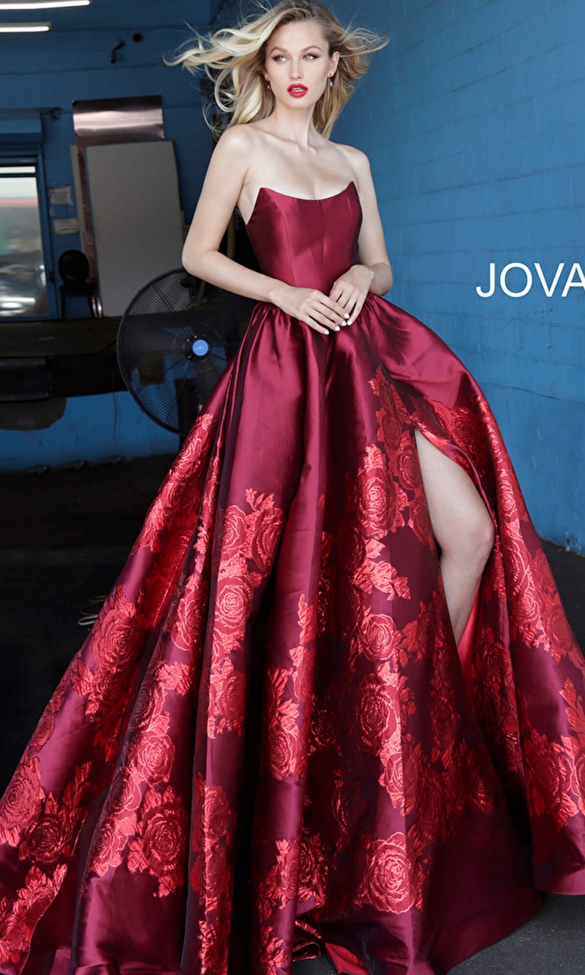 A woman poses indoors against a blue wall wearing the Jovani Strapless Floral Long Prom Ball Gown 02038, a floor-length red evening dress with a floral print and high slit.